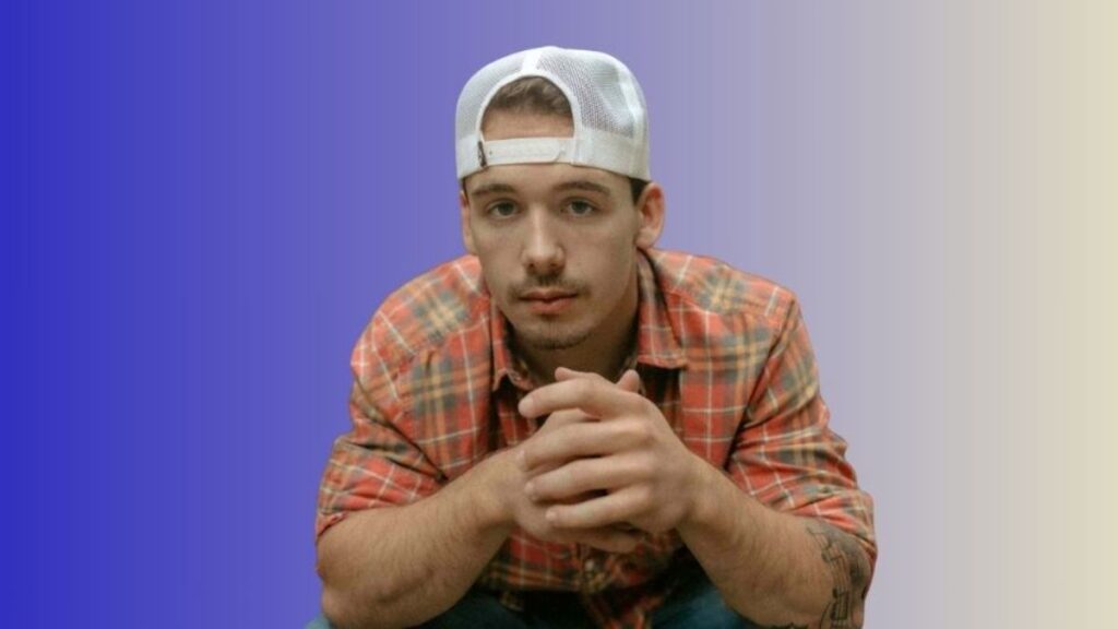 Noah Thompson net worth — country music artist wearing an orange plaid shirt and white cap, sitting with hands clasped against a blue gradient background.